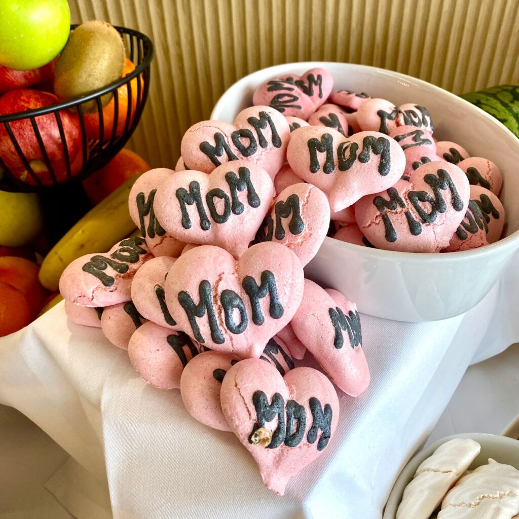 Bonbons en forme de cœurs roses ornés du mot "MOM", disposés dans un bol et sur une nappe blanche, accompagnés de fruits frais à côté.