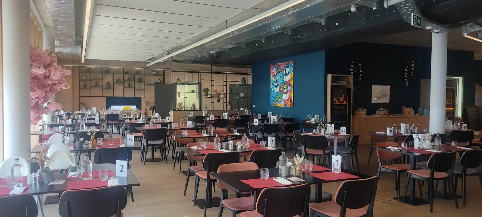 Salle de brunch conviviale avec tables dressées, chaises noires et sets rouges, ambiance chaleureuse et espace de jeux en fond.